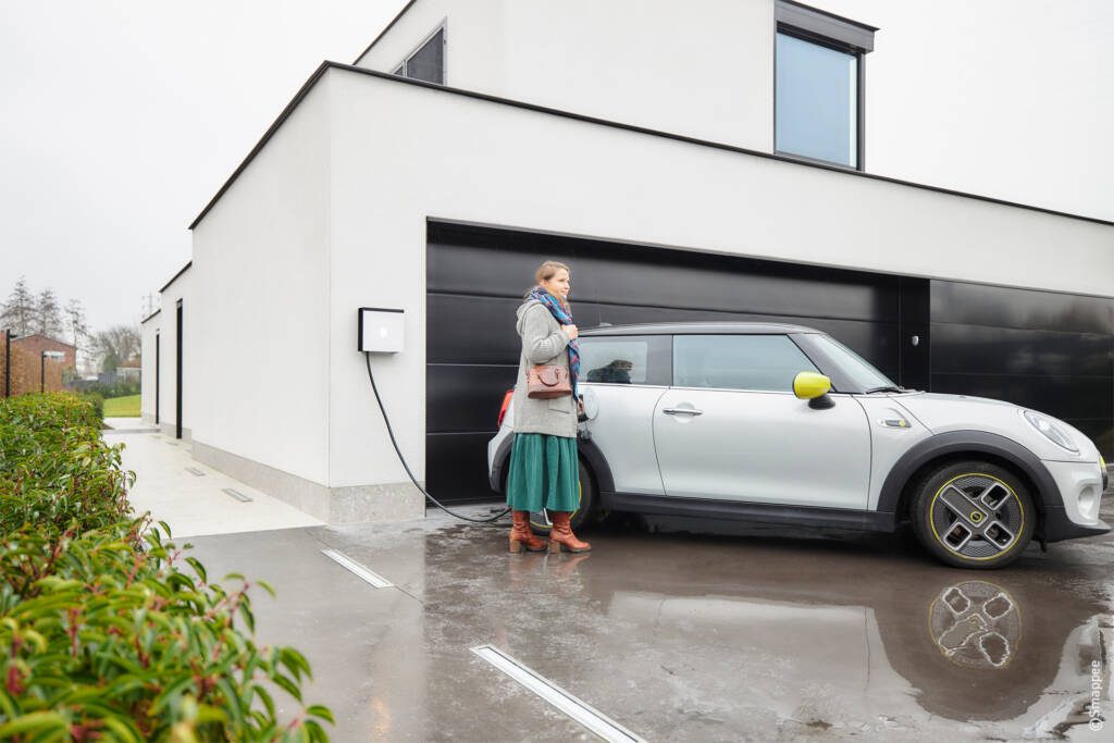 Een vrouw staat voor een huis met een auto ervoor geparkeerd, naast laadpalen.
