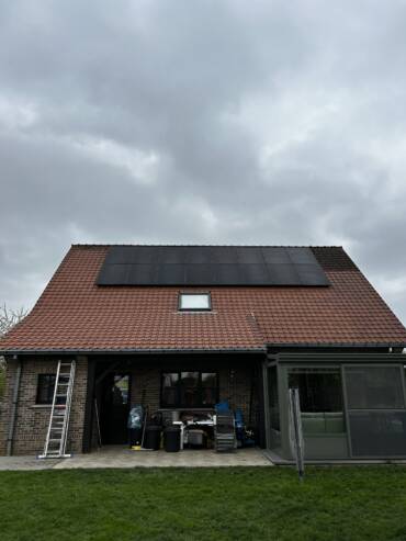 Une maison avec des tuiles rouges et des panneaux solaires est représentée sous un ciel nuageux. Une échelle repose contre le côté gauche et divers objets sont rangés sous le surplomb. Une petite structure vitrée se trouve à droite.