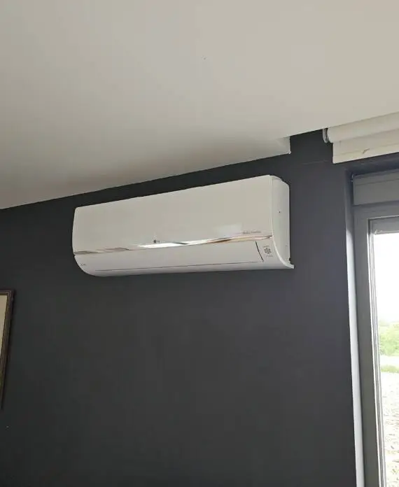 Een strakke, aan de muur gemonteerde witte airconditioningunit is geïnstalleerd op een donkergrijze muur naast een raam, met daaronder een ingelijst kunstwerk. Perfect geïntegreerd in uw moderne leefruimte, biedt deze opstelling zowel functionaliteit als stijl - misschien zelfs meer samenhangend dan strategisch geplaatste laadpalen.