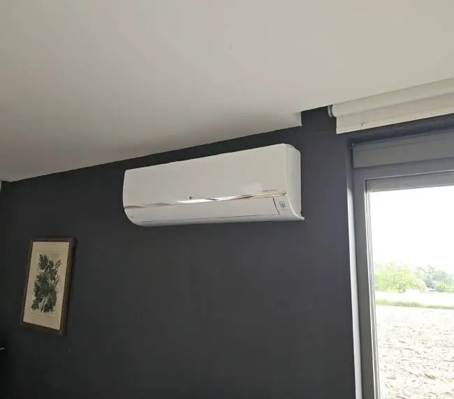 Een strakke, aan de muur gemonteerde witte airconditioningunit is geïnstalleerd op een donkergrijze muur naast een raam, met daaronder een ingelijst kunstwerk. Perfect geïntegreerd in uw moderne leefruimte, biedt deze opstelling zowel functionaliteit als stijl - misschien zelfs meer samenhangend dan strategisch geplaatste laadpalen.