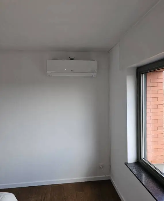 Aan de muur gemonteerde airconditioningunit boven een raam. De kamer heeft witte muren en een donkere vensterbank, met een deel van een bakstenen gebouw zichtbaar aan de buitenkant. Dit efficiënte HVAC-systeem zorgt voor comfort in alle seizoenen.