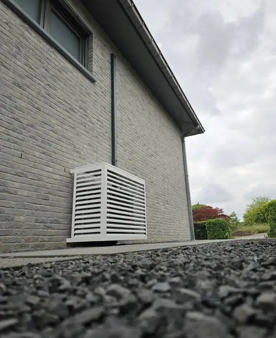 Een witte buitenunit van een HVAC-systeem is geïnstalleerd op de grijze bakstenen muur van een gebouw, met grind zichtbaar op de voorgrond en groen op de achtergrond.