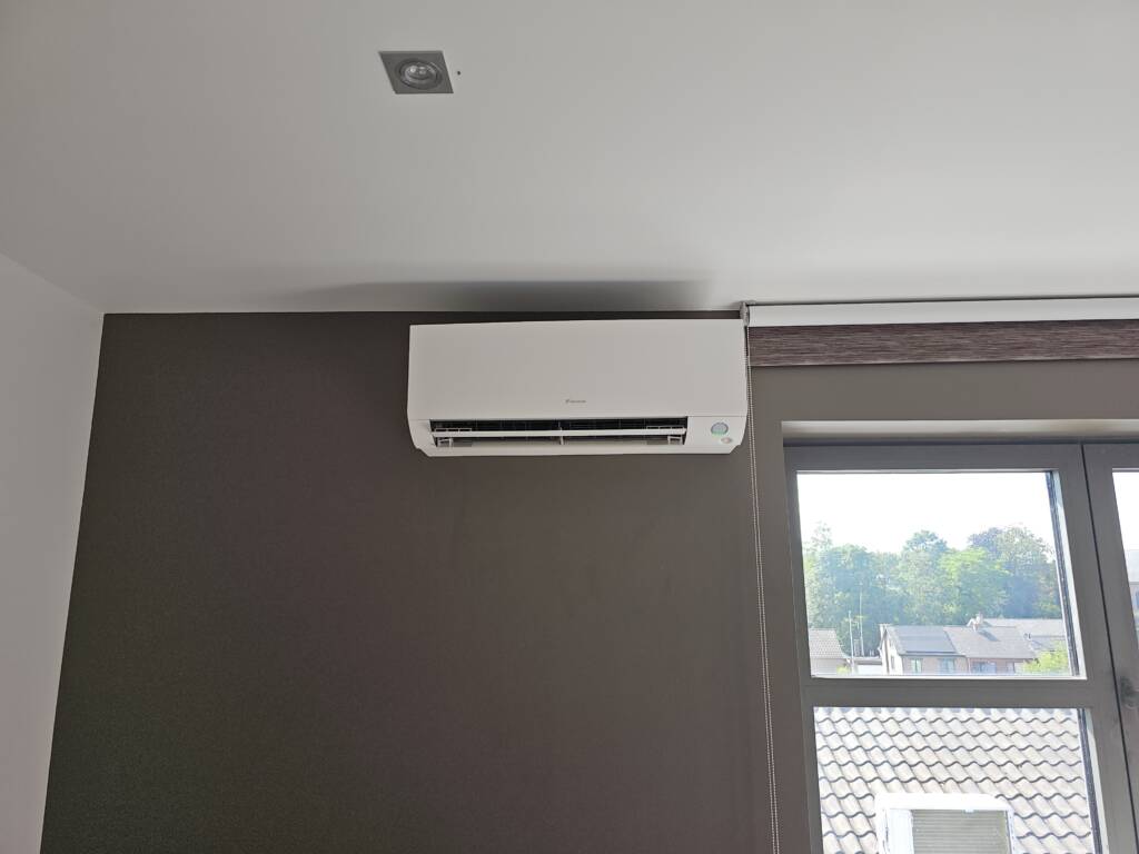 Een aan de muur gemonteerde airco-unit wordt boven een raam met een gedeeltelijk gesloten gordijn in een kamer met een donkere muur geïnstalleerd en zorgt voor een efficiënt koelsysteem.
