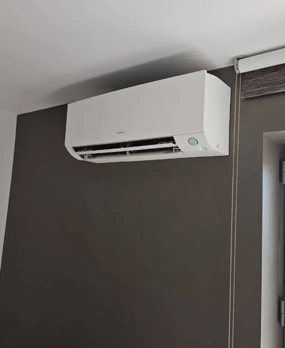 Aan de muur gemonteerde airconditioningunit boven een raam met witte afwerking en een uitschuifbaar rolgordijn, contrasterend met een donkere muur, zorgt voor een optimale klimaatregeling.