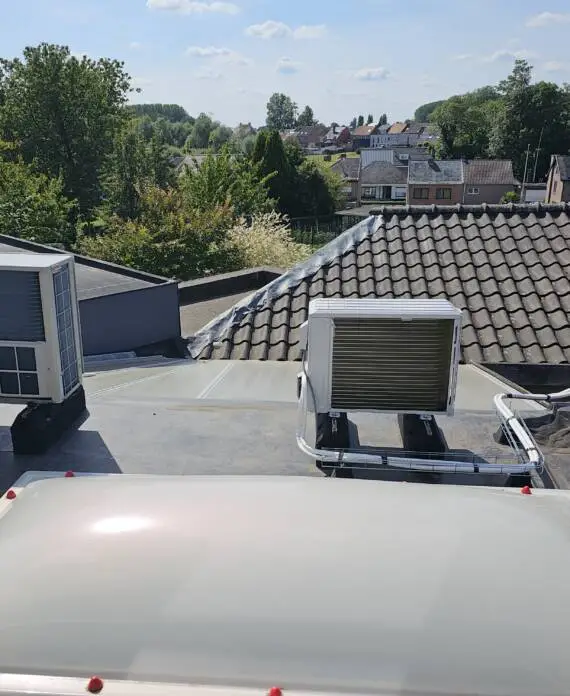 Twee HVAC-units zijn geïnstalleerd op een plat dak. De omgeving bestaat uit aangrenzende huizen, bomen en een heldere lucht met verspreide wolken. Deze airconditioningsystemen zorgen voor een optimale klimaatbeheersing en behouden het comfort voor de bewoners beneden.