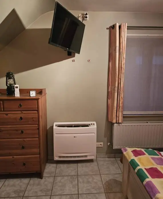 Een kamer met een aan de muur gemonteerde tv, een houten ladekast met een lantaarn en een klok, airconditioning, een raam met gordijnen en een deel van een bed met een kleurrijke deken.