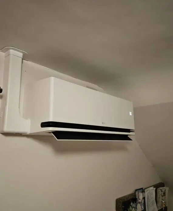 Aan de muur gemonteerde airconditioningunit in een hoek boven een raam, waarbij de gordijnen gedeeltelijk zichtbaar zijn.