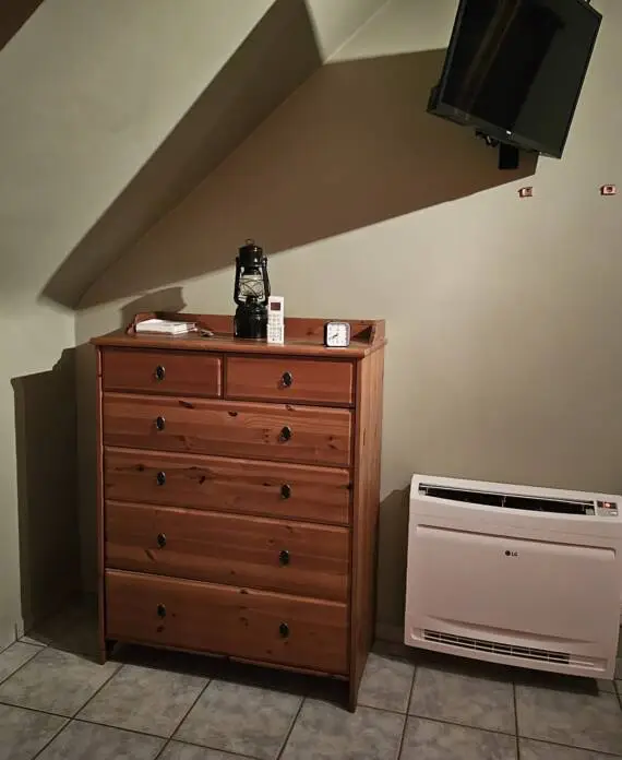 Houten dressoir met spullen erbovenop, naast een tv aan de muur en een kachel eronder. Kamer heeft schuin plafond en betegelde vloer.