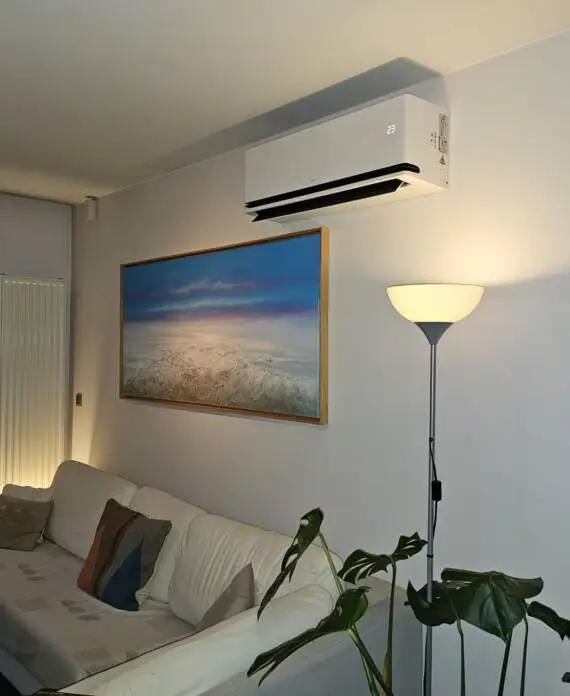 Woonkamer met een witte bank, een staande lamp en een aan de muur gemonteerde airconditioner. Een groot ingelijst zeegezicht hangt boven de bank. Gordijnen zijn gedeeltelijk dicht.