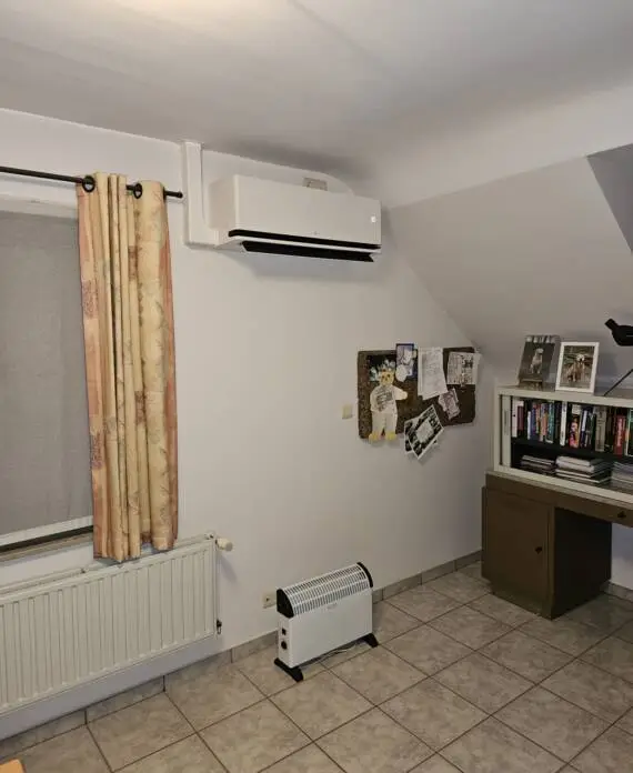 Kamer met airconditioner, radiator en ruimteverwarming. Bureau met boeken, een prikbord en gordijnen bedekken een raam. Tegelvloer.