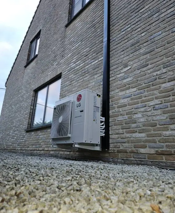 Een buitenairconditioningunit gemonteerd op de bakstenen muur van een gebouw, met twee zichtbare ramen. Een grindgrond is op de voorgrond.