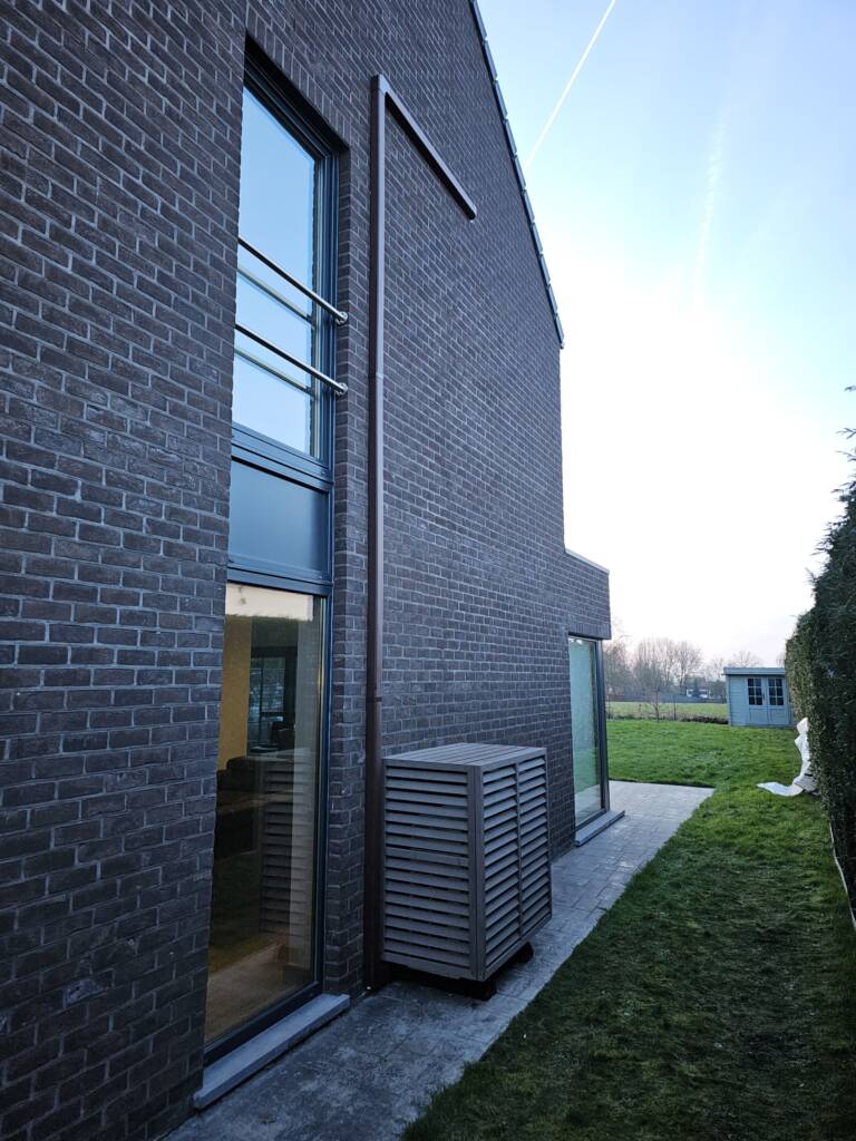 Modern bakstenen huis met grote ramen, een airconditioningunit met een lamellenafdekking en een grasveld met een schuur op de achtergrond.